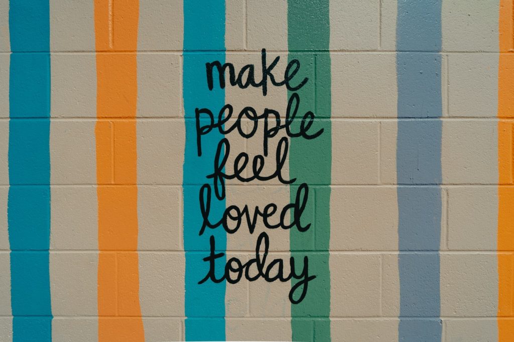 A stone wall with colored stripes from top the bottom and writing in the middle saying make people feel loved today.