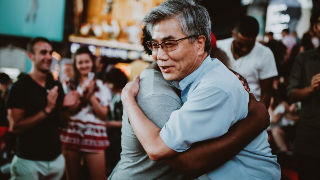Two people are hugging and you can see the happy for of a man hugging someone else while others in a crowd clap behind them